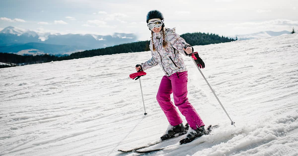 Skier practicing safe technique on a snowy mountain slope, demonstrating balance and control to help prevent ski injuries, with mountains and trees in the background under a clear winter sky.
