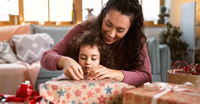 Woman in a red sweater wrapping holiday gifts at a table near a decorated Christmas tree, demonstrating proper posture and workspace setup for comfortable gift wrapping.