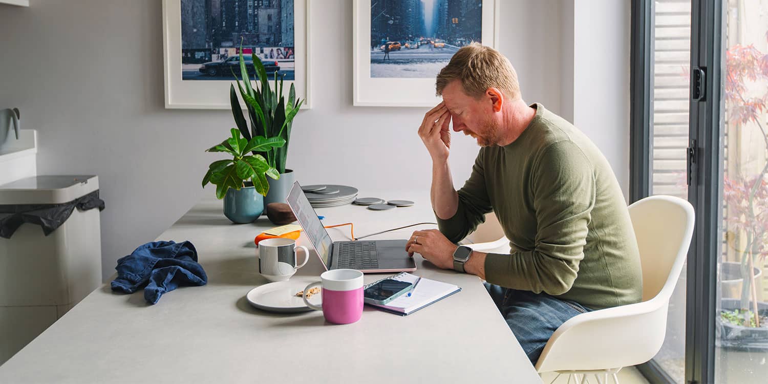 Man sitting at table with laptop holding head, showing stress and tension from work