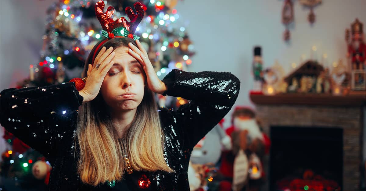 Woman in reindeer headband holding her head in front of a Christmas tree, showing holiday stress and the need for relief and balance