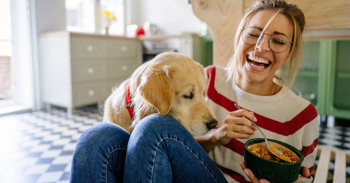 Young woman laughing on the kitchen floor with her dog while eating a healthy meal, illustrating joy and balance for a health and wellness checklist.