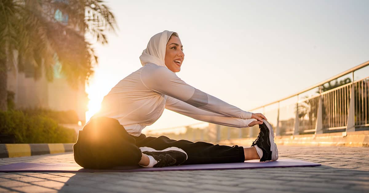 Woman in a hijab smiling as she stretches forward to touch her foot on a yoga mat at sunrise, illustrating how to move past common stretching myths and support flexible, comfortable movement.