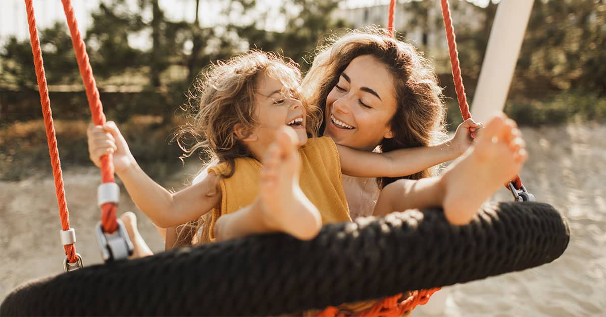 Mother laughing with her young daughter on an outdoor swing, representing how realistic new year's resolutions can support more energy, movement, and joyful everyday moments together.